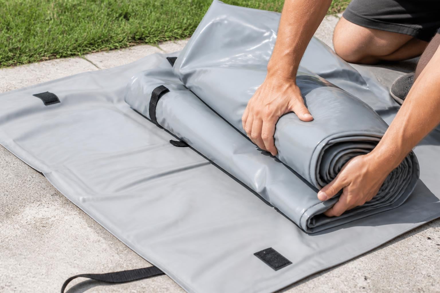 Close-up illustration of hands carefully folding a deflated gray inflatable boat on a driveway, rolling it toward a carr